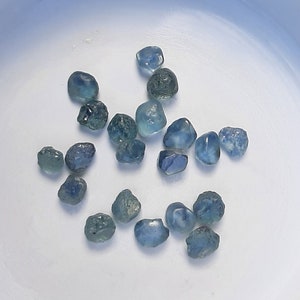 May include: A selection of small, rough blue gemstones, possibly sapphires or aquamarines, are arranged in a white, shallow bowl. The stones display varying shades of blue and appear to be natural, unpolished specimens.
