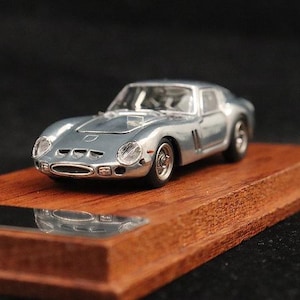 Puede incluir: Un coche modelo plateado con un diseño elegante, montado sobre una base de madera rectangular. El vehículo en miniatura presenta faros detallados, una toma de aire en el capó y un perfil bajo. La base mide unos 15 cm de largo.