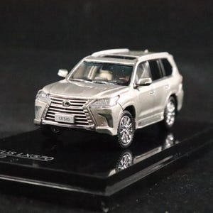 May include: A silver Lexus LX 570 model car on a black display stand. The miniature SUV has detailed features, including a sunroof and chrome accents. The model is a 1:1000 scale replica.