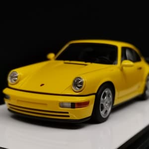 May include: A bright yellow sports car model with black accents, set against a dark background. The miniature vehicle features detailed headlights, wheels, and a sleek, aerodynamic design. The car is a collectible item.