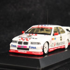 May include: A white race car model with red and black accents, featuring sponsor logos and the text "Hurststeiner" and "Fina". The car has a spoiler and is displayed on a black base.