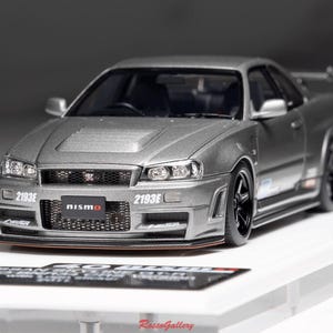 Könnte beinhalten: Ein silbernes Modellauto mit schwarzen Rädern und einem Heckspoiler. Das Auto trägt das Wort "nismo" auf der Vorderseite und die Nummer "2193E" auf der Vorder- und Seite. Das Auto steht auf einer weißen Oberfläche.