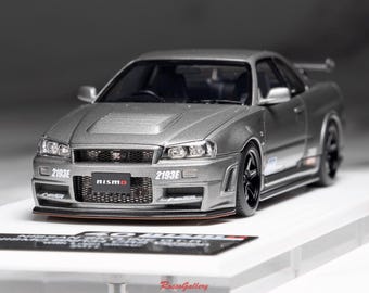 May include: A silver model car with black wheels and a rear spoiler. The car has the word "nismo" on the front and the number "2193E" on the front and side. The car is on a white surface.