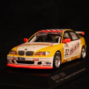 May include: A miniature BMW 320i race car model. The car is white and yellow with red accents, featuring the number 32. It has sponsor logos and a checkered flag design on the roof. The model sits on a black base.