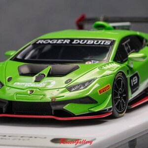 Puede incluir: Un modelo de coche de carreras Lamborghini Huracan de color verde brillante. El modelo presenta detalles en negro, el número 19 y logotipos de patrocinadores, incluido "ROGER DUBUIS" en el parabrisas. El coche tiene un alerón trasero y está sobre una superficie blanca.