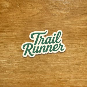May include: A white sticker featuring the words "Trail Runner" in a dark green, cursive font. The sticker has a white border and is set against a wooden background.