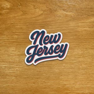 Può includere: Un adesivo con la scritta "New Jersey" in un carattere corsivo. Il testo è blu navy con un contorno rosso e un bordo bianco. L'adesivo è su una superficie di legno.