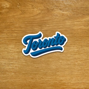 May include: A blue and white sticker featuring the word "Toronto" in a stylized script font. The word is in a dark blue colour with a white outline and a lighter blue shadow, set against a wooden background.