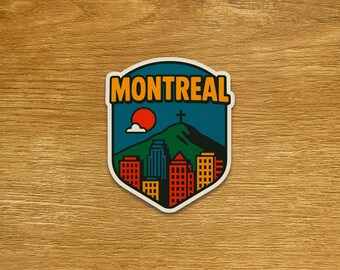 May include: A sticker with a shield design featuring the word "MONTREAL" in orange letters. The design includes a green mountain, a red sun, and a cityscape with red, blue, and yellow buildings. A cross is atop the mountain.