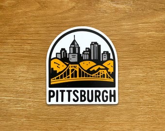 Op de afbeelding: Een witte sticker met een zwart-gele illustratie van de skyline van Pittsburgh en een brug. Het woord "PITTSBURGH" staat in het zwart onderaan de sticker.