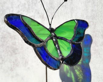 May include: A stained glass butterfly ornament featuring vibrant green and deep blue glass panels held together by dark metal solder lines. The butterfly has two thin metal antennae and is mounted on a slender metal rod for display purposes.