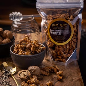 May include: A clear plastic bag filled with honey glazed walnuts. The bag is labeled "DeM' Nutz" and "Honey Glazed Walnuts". The walnuts are a light brown color and have a shiny glaze. There is a bowl of walnuts in the background.
