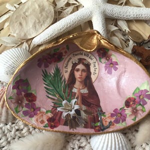 May include: A decorative seashell with a gold-painted rim and a pink interior. The shell features a central image of a saint surrounded by floral and botanical elements. The text on the image reads "St. Philomena Powerful with God - Pray for us."