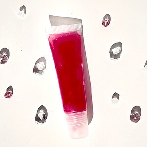 Könnte beinhalten: Ein durchsichtiges Plastikröhrchen mit rosa Lipgloss. Das Röhrchen hat einen weißen Deckel und steht auf einer weißen Oberfläche.