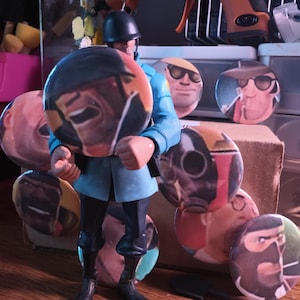 May include: A figurine of a soldier in a blue uniform and black boots, holding several round buttons with character portraits. The buttons feature various faces and expressions. The background includes storage containers and art supplies.