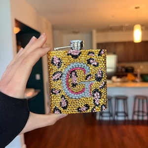 Puede incluir: Una petaca de metal rectangular cubierta con pequeños diamantes de imitación brillantes dispuestos en un patrón de leopardo en tonos oro, negro y rosa. El centro presenta una letra G grande y estilizada, delineada en plata y rellena de piedras rosas.