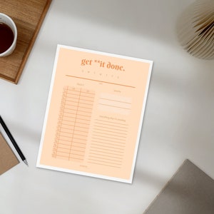 May include: A peach-colored daily planner with the text "get **it done." A cup of tea, a pencil, and a notepad are on the table. The planner has sections for the date, tasks, and priorities.