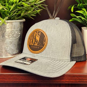 May include: A gray trucker hat with a black mesh back. The hat features a circular leather patch with the words "Sequoia National Park" and a graphic of trees. The hat has a curved brim and a Richardson 112 tag.