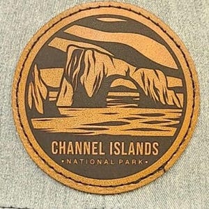 May include: A round, brown leather patch featuring a landscape design with the text "CHANNEL ISLANDS NATIONAL PARK." The design depicts a rock formation over water. The patch has a stitched border.