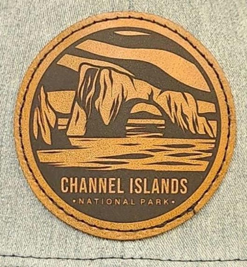 May include: A round, brown leather patch featuring a landscape design with the text "CHANNEL ISLANDS NATIONAL PARK." The design depicts a rock formation over water. The patch has a stitched border.