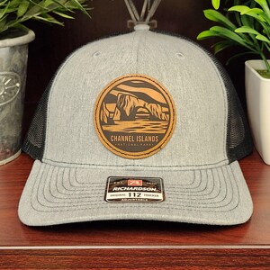 May include: Gray trucker hat with a black mesh back and a curved brim. The hat features a circular patch with the text "CHANNEL ISLANDS NATIONAL PARK" and an illustration of rock formations. The hat also has a "RICHARDSON" tag.