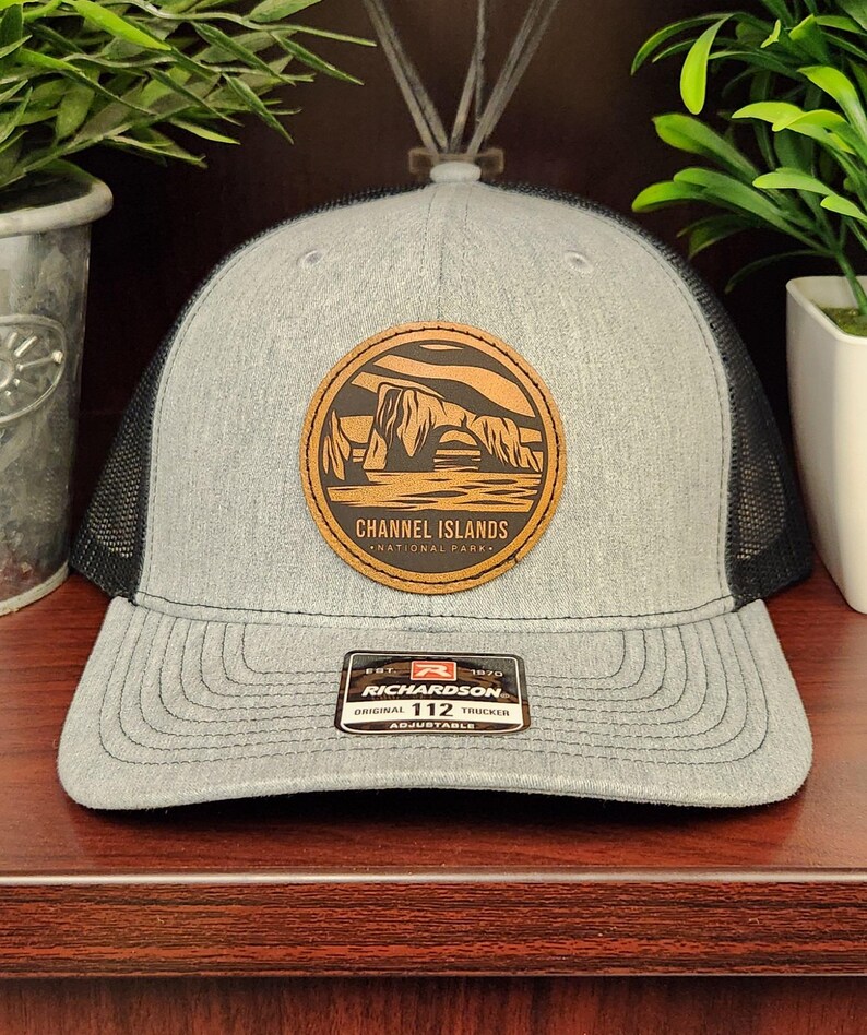 May include: Gray trucker hat with a black mesh back and a curved brim. The hat features a circular patch with the text "CHANNEL ISLANDS NATIONAL PARK" and an illustration of rock formations. The hat also has a "RICHARDSON" tag.
