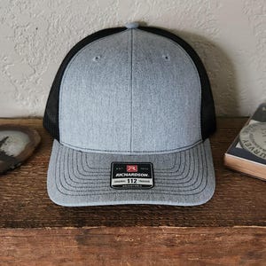 May include: A gray and black trucker hat with a curved brim. The front panel is gray, and the back is black mesh. The hat has a patch that reads "RICHARDSON" and "112 Trucker". Adjustable.