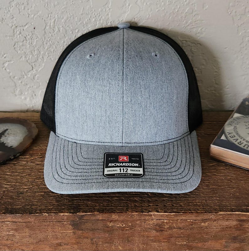 May include: A gray and black trucker hat with a curved brim. The front panel is gray, and the back is black mesh. The hat has a patch that reads "RICHARDSON" and "112 Trucker". Adjustable.