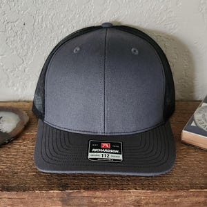 May include: A gray and black trucker hat with a curved brim. The front panel is gray, and the mesh back is black. The hat features a Richardson 112 patch on the front and is adjustable.