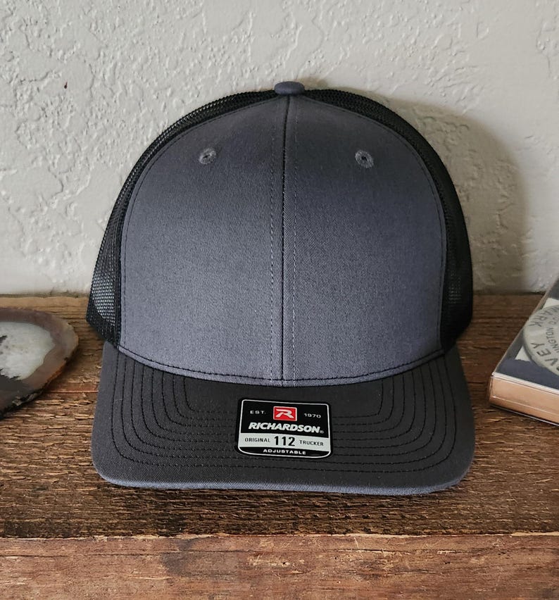 May include: A gray and black trucker hat with a curved brim. The front panel is gray, and the mesh back is black. The hat features a Richardson 112 patch on the front and is adjustable.