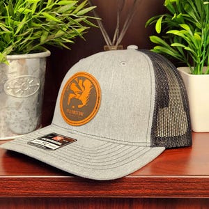 May include: Gray trucker hat with a black mesh back and a curved brim. The hat features a circular patch with an orange graphic and the word "Yellowstone". The hat is displayed on a wooden surface.
