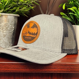May include: A gray trucker hat with a black mesh back and a curved brim. The hat features a round, brown leather patch with an orange landscape design and the word "VOYAGEURS".