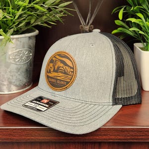 May include: A gray trucker hat with a black mesh back and a curved brim. The hat features a round leather patch with a landscape design and the text "Channel Islands National Park". A "Richardson" tag is visible.