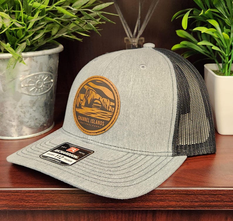 May include: A gray trucker hat with a black mesh back and a curved brim. The hat features a round leather patch with a landscape design and the text "Channel Islands National Park". A "Richardson" tag is visible.