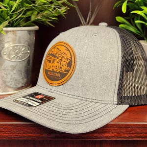 May include: A gray baseball cap with a black mesh back and a curved brim. The cap features a circular leather patch with the words "Mesa Verde National Park" and an illustration of a cliff dwelling. The cap has a Richardson 112 tag.