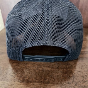 May include: A dark gray trucker hat with a mesh back and a solid front. The hat has a curved brim and an adjustable snap closure. The hat is displayed on a wooden surface.