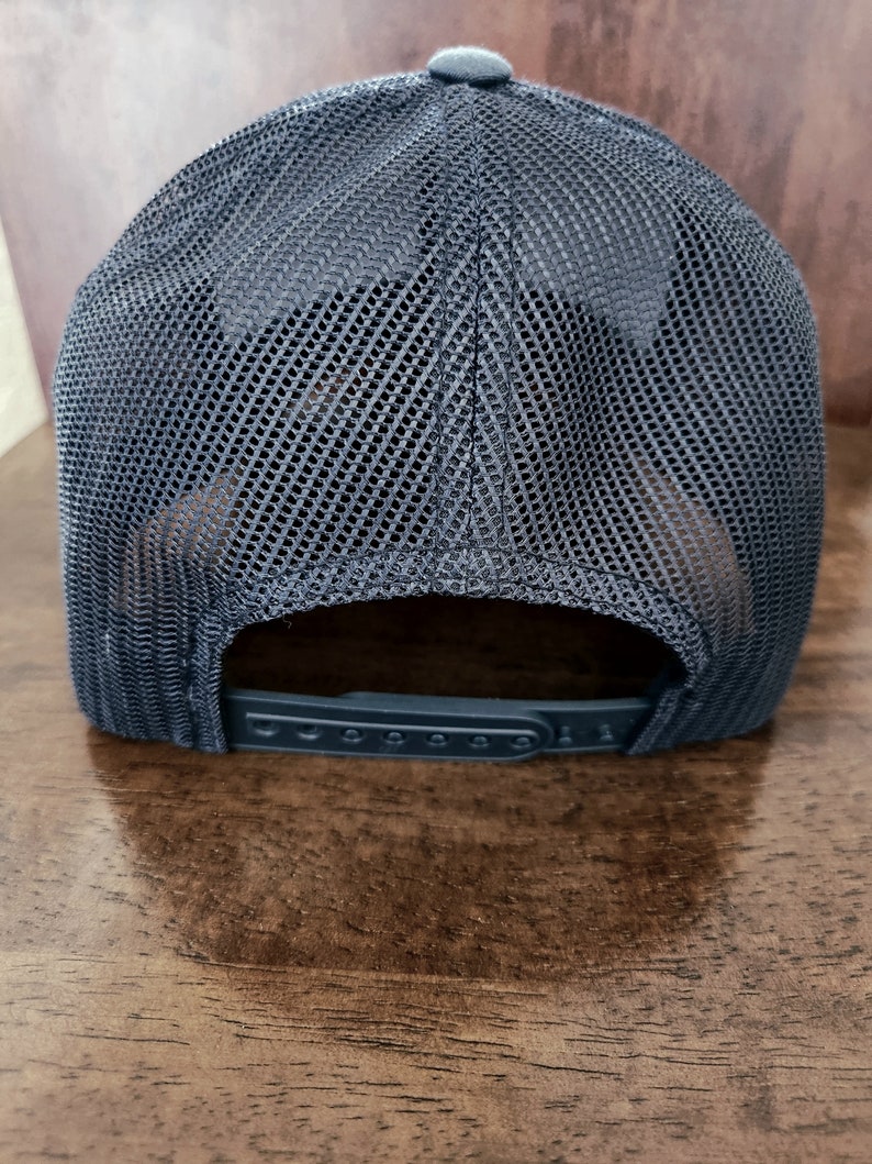 May include: A dark gray trucker hat with a mesh back and a solid front. The hat has a curved brim and an adjustable snap closure. The hat is displayed on a wooden surface.