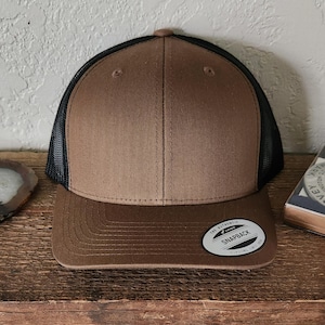 May include: A brown and black trucker hat with a curved brim. The hat features a brown front panel and brim, a black mesh back, and a snapback closure. A small, circular tag on the brim reads "The Authentic Snapback".