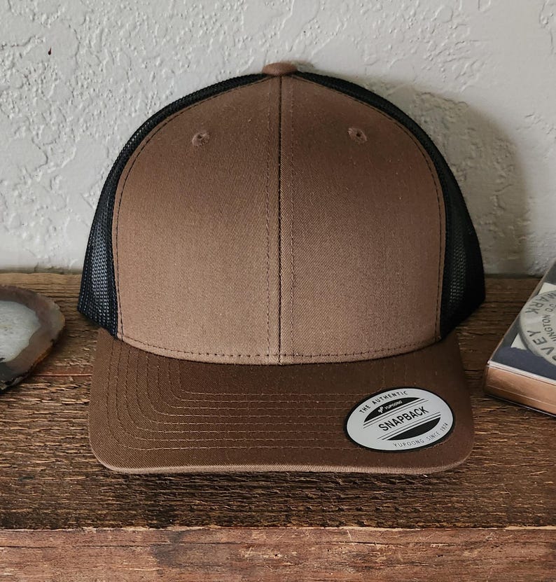 May include: A brown and black trucker hat with a curved brim. The hat features a brown front panel and brim, a black mesh back, and a snapback closure. A small, circular tag on the brim reads "The Authentic Snapback".