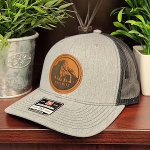 May include: Gray trucker hat with a black mesh back and a brown leather patch featuring a mountain silhouette and the text "Yosemite National Park". The hat has a curved brim and an adjustable strap.