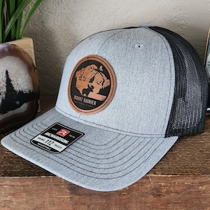 May include: A gray and black trucker hat with a brown leather patch featuring a silhouette of a mountain range and the text "Mount Rainier National Park". The hat is made by Richardson 112.