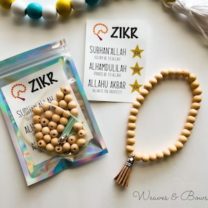 May include: A Zikr prayer bead set with wooden beads, a tassel, and a card with text. The card reads "Zikr" and lists phrases in Arabic with English translations. A beaded garland with white, yellow, and teal beads is also present.
