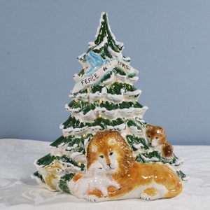May include: A ceramic Christmas tree with a green and white snowy design. A lion and lamb are nestled at the base, with a fox and rabbit nearby. A banner reads "Peace on Earth" with a blue dove.