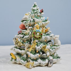 May include: A ceramic Christmas tree with a snowy effect, adorned with various woodland creatures and a festive wreath. The tree features a variety of animals, including an owl, raccoon, birds, and squirrels. The tree is lit with small, warm lights.