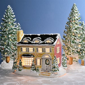 May include: A ceramic Christmas village house with a red and yellow facade, black roof, and glowing windows. Two snow-covered Christmas trees flank the house. The scene is set on a snowy base, creating a festive holiday display.