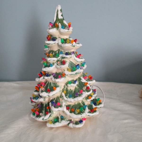 May include: A ceramic Christmas tree with a cream-colored base, green branches, and colorful light bulbs. The tree is decorated with white snow accents. A white electrical cord extends from the back of the tree.