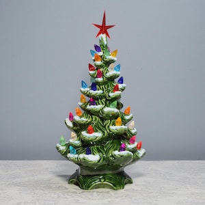 May include: A green ceramic Christmas tree with a red star on top and multi-colored lights. The tree is covered in white faux snow.