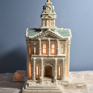 May include: A white ceramic miniature county courthouse with a green roof and a clock tower. The building has a sign that reads "County Court House". The windows are lit up, creating a warm glow.