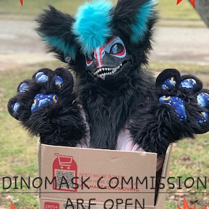 Op de afbeelding: Een zwart en blauw dinosaurus-fursuit met een tandenrij, zittend in een kartonnen doos met de tekst "Dino Mask Commission Are Open" erop gedrukt.