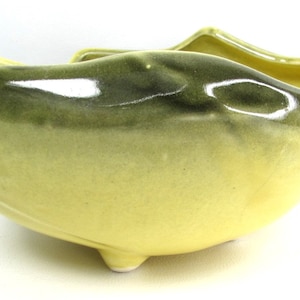 Pode incluir: Um vaso de cerâmica em forma de peixe amarelo e verde com acabamento esmaltado. O vaso tem uma boca larga e aberta e uma cauda que se enrola para cima. Tem três pequenas patas na parte inferior.